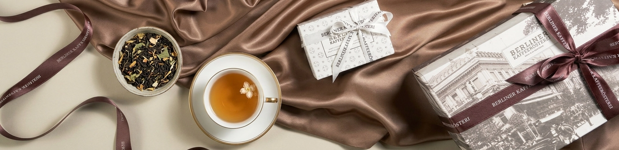 Elegante Geschenkboxen mit Tee auf schimmerndem Satinstoff arrangiert – daneben eine Tasse goldener Tee und lose Teeblätter in stilvoller Premium-Ästhetik. - Hintergrund