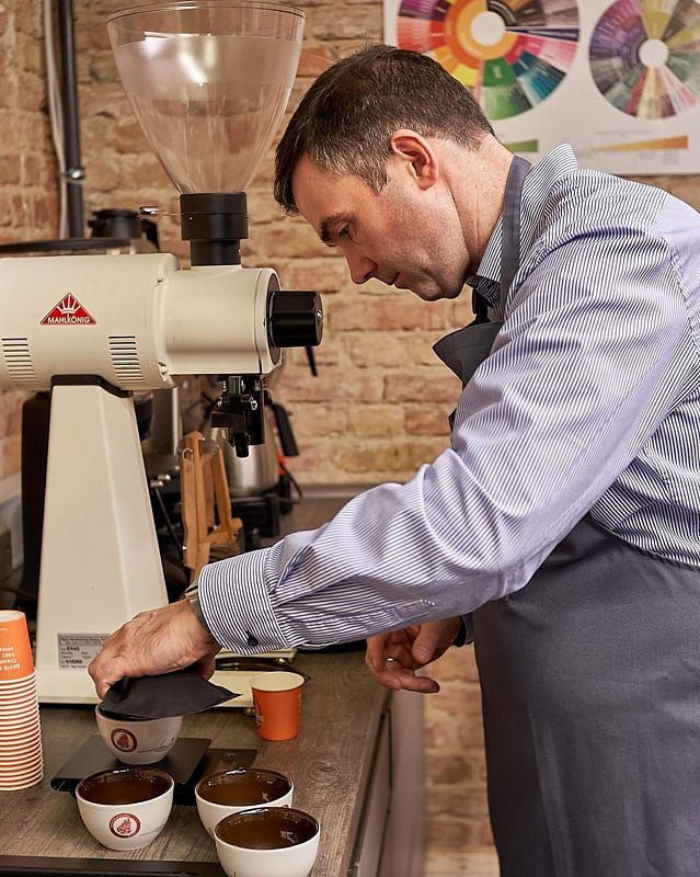 Ein Mann bewertet frisch gebrühten Kaffee beim Cupping, indem er mehrere Tassen neben einer professionellen Kaffeemühle auf einem Tisch prüft.