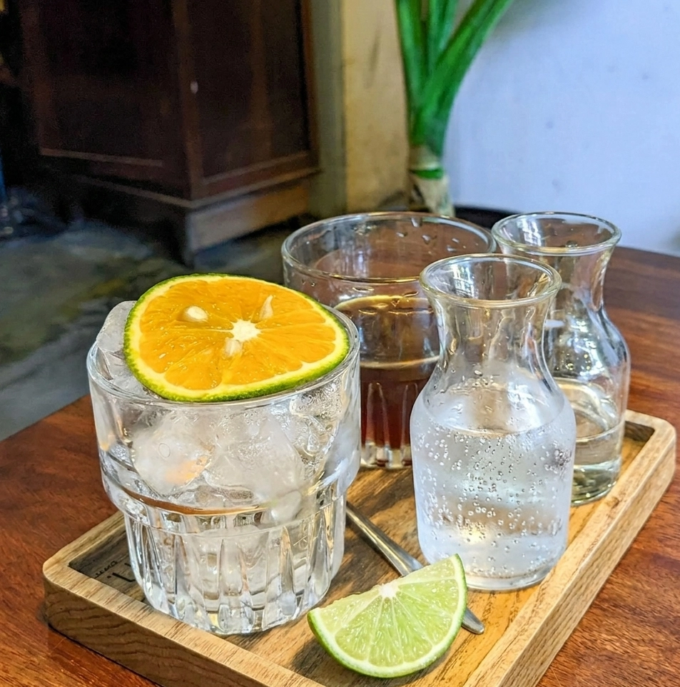 Sparkling Lime Americano Serving Set auf Holztablett: Eisglas mit Orangenscheibe, kleine Karaffe mit Sprudelwasser, Glas mit Espresso und Limettenspalte als Garnitur.